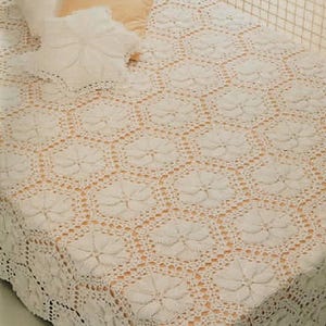 Puede incluir: Colcha de ganchillo color crema con un patrón floral. La colcha está drapeada sobre una cama, con almohadas visibles. El trabajo de ganchillo presenta un diseño floral repetido y los bordes son festoneados.