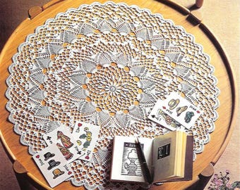 Può includere: Un centrino bianco all'uncinetto con un intricato motivo floreale, appoggiato su un tavolo di legno. Carte da gioco e un piccolo libro aperto con una penna sono disposti attorno al centrino, creando un'estetica vintage e accogliente.