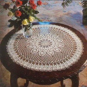 May include: A round, off-white crocheted doily featuring an intricate, radiating floral pattern with a scalloped edge. The doily is centered on a dark brown wooden table, accompanied by a clear glass vase containing red and yellow roses.