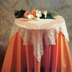 May include: A round table covered with an orange tablecloth and a white, crocheted lace doily. The doily has a scalloped edge and a floral pattern. Fresh flowers, including orange and white blooms, are arranged on top.