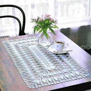 Könnte beinhalten: Ein weißes, rechteckiges, gehäkeltes Deckchen mit detailliertem Lochmuster liegt auf einem Holztisch. Eine klare Glasvase mit rosa Blumen und eine weiße Kaffeetasse stehen ebenfalls auf dem Tisch. Im Hintergrund ist ein schwarzer Stuhl zu sehen.