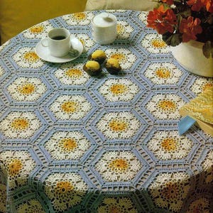 以下が含まれることがあります： 繰り返し六角形の模様が特徴の、丸いかぎ針編みのテーブルクロス。白、黄色、水色のデザインです。コーヒーカップ、砂糖入れ、お菓子がテーブルに置かれ、赤い花の入った花瓶も置かれています。