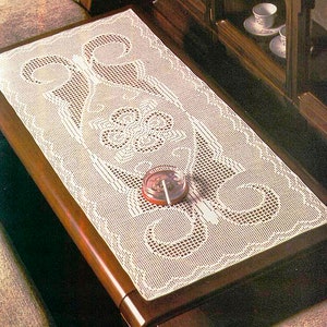 May include: White crocheted table runner with a delicate floral design. The runner is laid on a wooden table with a small ashtray on top.