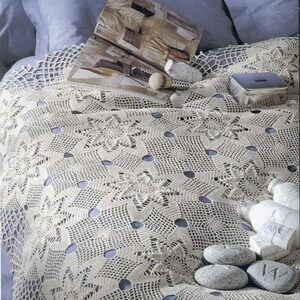 Pode incluir: Um colcha de crochê creme com um padrão floral e geométrico. A colcha está sobre uma cama com travesseiros azuis. Uma revista, uma pequena caixa de prata e pedras decorativas estão sobre a colcha.