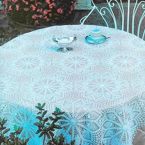 以下が含まれることがあります： 白いかぎ針編みのレースのテーブルクロスが丸いテーブルを覆っています。複雑な模様が見え、小さな銀色の皿とティーカップとソーサーが上に置かれています。テーブルは屋外にあり、背景には白い金属製の椅子があります。