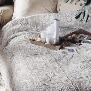 May include: A cream-colored, floral-patterned bedspread covers a bed. A wooden tray holds white mugs, a vase, and pastries. Small framed pictures and decorative items are also on the tray. The bedspread's design features repeating floral motifs.
