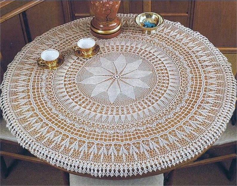 Puede incluir: Mantel de mesa de crochet blanco con un delicado dise&ntilde;o floral en el centro. El mantel est&aacute; puesto sobre una mesa de madera con dos tazas de t&eacute; blancas y un taz&oacute;n peque&ntilde;o.