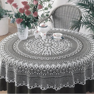 Puede incluir: Un mantel de crochet blanco con un delicado patrón de encaje, que presenta un diseño circular en el centro. El mantel está puesto sobre una mesa marrón oscuro con una taza de té y un platillo blancos encima.