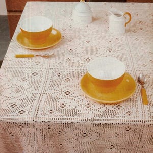 May include: A crocheted tablecloth with a floral pattern covers a table set for tea. Two yellow teacups and saucers, spoons, a sugar bowl, and a creamer are visible. The tablecloth is a cream color.