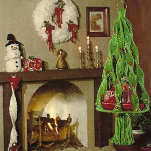 May include: A green crocheted Christmas tree ornament hanging from a ceiling hook. The tree is decorated with red ornaments and small gifts. The tree is hanging in front of a fireplace with a lit fire.