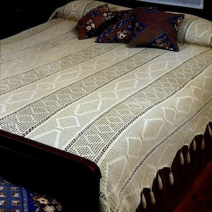 Puede incluir: Una colcha de ganchillo de color crema con un patrón geométrico y un borde con flecos. La cama está hecha con cojines decorativos en tonos marrones y azules. Un cojín a juego es visible en el lateral de la cama.