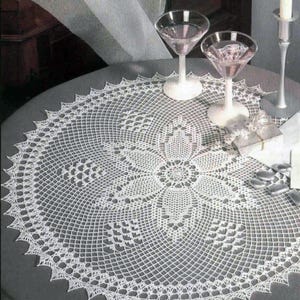 Puede incluir: Un tapete blanco de ganchillo con un diseño floral central y un borde festoneado. Dos copas de martini y un candelabro están sobre la mesa. El tapete es circular y cubre la mayor parte de la superficie de la mesa.