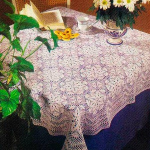 Puede incluir: Un mantel blanco, intrincadamente tejido a ganchillo, con un borde festoneado cubre una mesa. Un jarrón con flores blancas, una taza de té y un libro están sobre la mesa. Plantas verdes en primer plano.