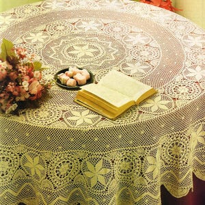 Könnte beinhalten: Ein cremefarbenes, rundes gehäkeltes Tischtuch mit floralem und geometrischem Muster. Das Tischtuch ist über einem Tisch drapiert, mit einem offenen Buch und einem kleinen Teller mit rosa Süßigkeiten. Ein Blumenstrauß befindet sich links.