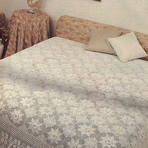 May include: A white crocheted bedspread featuring a repeating floral motif pattern with intricate openwork details and a scalloped edge. The bedspread covers a bed with a tan patterned headboard and two square throw pillows in beige and white.