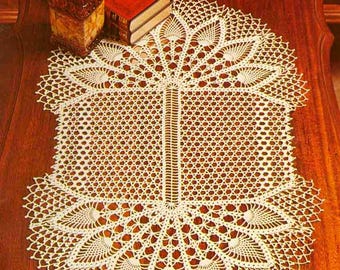 Può includere: Tovaglietta all'uncinetto fatta a mano, color crema, con un design traforato dettagliato. La tovaglietta presenta un pannello centrale verticale e motivi a forma di ventaglio sui bordi. Posizionata su una superficie di legno scuro, con un libro rosso e una lampada decorativa sullo sfondo.