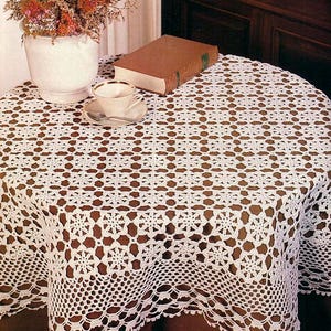 May include: A white, crocheted tablecloth with a detailed floral pattern covers a round table. A teacup and saucer, a book, and a vase of flowers are on the table. The tablecloth has a scalloped edge.