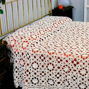 May include: A white, open-weave crochet bedspread with a repeating circular pattern, covering a bed with a gold-colored metal frame. The bedspread has a scalloped edge and is set against a red background.