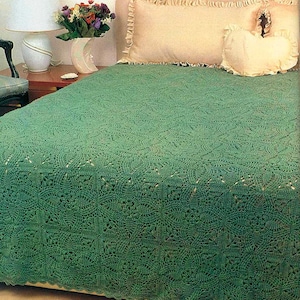 May include: A handmade, emerald green crochet bedspread with a detailed floral pattern. The bedspread covers a bed with cream-colored pillows and a headboard. A lamp and vase of flowers are visible on a nightstand.