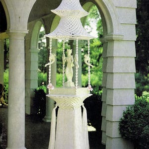May include: A white macramé hanging structure titled Macramé Enchantment Book 4, featuring a pagoda-style top, a central figurine, and four twisted support columns. The base includes long white fringe, and the text Fountain of the Orient is visible.