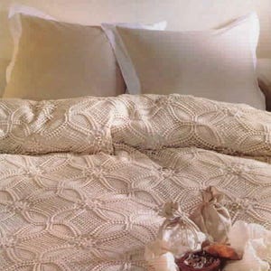 May include: A cream-colored crochet bedspread featuring a repeating circular and floral pattern. The bedspread is draped over a bed with two beige pillows and two white pillows. In the foreground, two small fabric pouches sit on the bedspread.