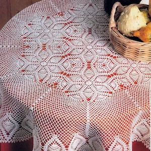 Pode incluir: Uma toalha de mesa redonda branca de crochê com um design detalhado em renda. O padrão intrincado cobre toda a superfície, com uma borda ondulada. Uma cesta com cabaças e um pote de cerâmica são visíveis no fundo.