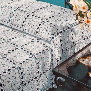 Pode incluir: Uma colcha branca com um padrão de crochê intrincado e repetitivo, com pequenos motivos florais escuros de quatro pétalas integrados no design. O tecido cobre uma cama, exibindo uma construção de renda texturizada e vazada.