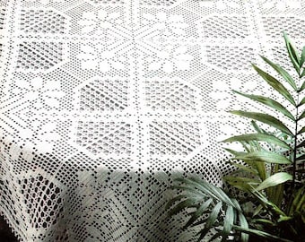 Può includere: Una tovaglia bianca, finemente lavorata all'uncinetto con un motivo geometrico, copre un tavolo rotondo. Il delicato design in pizzo è visibile, con una composizione floreale e una tazza da tè che aggiungono un tocco di eleganza. Una pianta con foglie verdi è in primo piano.