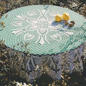 Pode incluir: Uma mesa redonda coberta com uma toalha de mesa de crochê branca e verde. Dois limões amarelos e três nozes castanhas estão sobre a mesa. A toalha de mesa tem um design detalhado e vazado com uma borda ondulada.