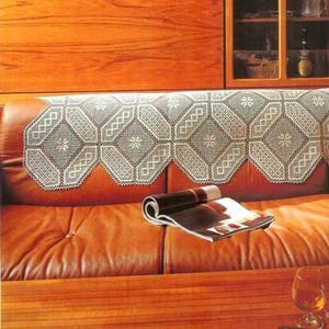 May include: A decorative, white lace doily with a geometric pattern is draped over the back of a brown leather couch. A rolled-up magazine rests on the couch. The scene is set in a room with wooden furniture.