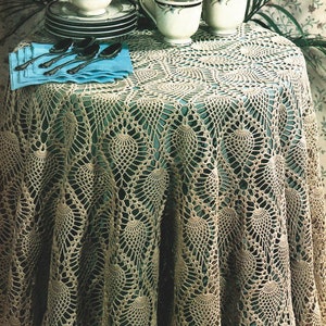 Peut inclure: Une nappe de table en crochet avec un motif délicat et complexe d'ananas. La nappe est beige et a un bord festonné. Elle est posée sur une table verte et a une serviette bleue et des cuillères en argent sur le dessus.