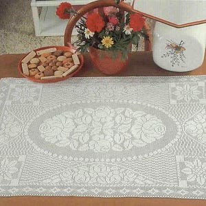 Pode incluir: Uma toalha de mesa retangular branca de crochê com um design floral. O oval central apresenta rosas, rodeado por padrões detalhados e motivos florais nos cantos. A toalha de mesa está colocada sobre uma mesa de madeira.