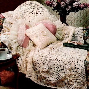 Puede incluir: Una manta de ganchillo color crema con motivos florales cubre un sofá estampado floral. La imagen incluye cojines a juego y una taza de café en una mesa auxiliar, creando una escena cálida y acogedora. El diseño de la manta es calado.