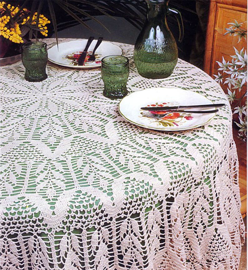 May include: A white crocheted tablecloth with a delicate floral pattern, laid over a green table with two plates, glasses, and a pitcher.