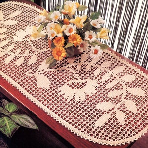 May include: A crocheted oval table runner with a delicate floral pattern in a cream color. The runner is laid on a wooden table with a bouquet of yellow and white flowers in a vase.