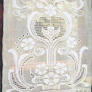 May include: Cream-colored, open-weave crochet curtain with a floral and scroll design. The curtain is hung on a wooden rod and features a detailed pattern of flowers, leaves, and decorative elements. The bottom edge has a scalloped trim.