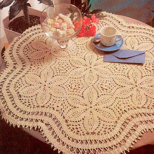 May include: A round, cream-colored crochet tablecloth featuring an intricate floral pattern with a scalloped edge. The textile is crafted with fine yarn, displaying a detailed openwork design that radiates from the center to the border.