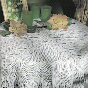 May include: White crocheted table cloth with a delicate lace pattern. The tablecloth is laid on a table with a green background and features a floral design in the center.