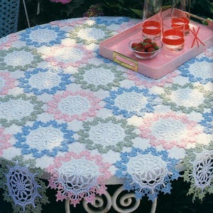 May include: A crocheted table cloth with a white background and a repeating pattern of blue, pink, and green flowers. The tablecloth is on a white table with a pink tray and glasses on top.