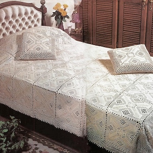 May include: A cream-colored crocheted bedspread and two matching square throw pillows featuring an intricate geometric pattern of interconnected diamonds and squares. The textile set is draped over a bed with a tufted headboard and wooden frame.