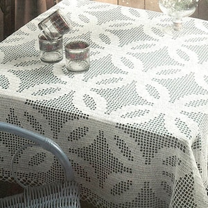 Pode incluir: Toalha de mesa de crochê branca com um delicado padrão floral. A toalha de mesa está posta sobre uma mesa com três copos de vidro transparentes e um vaso de flores brancas.