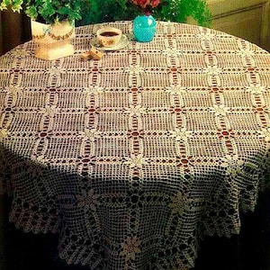 Puede incluir: Un mantel redondo de ganchillo en color blanco roto con un patrón de rejilla geométrico repetitivo y motivos florales. El intrincado diseño de encaje cubre toda la superficie, cayendo sobre los bordes de una mesa circular con laborioso detalle.