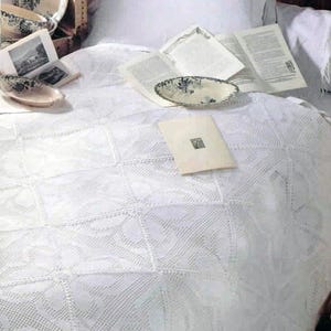 May include: A white crocheted bedspread featuring a repeating diamond and floral pattern. The bedspread is layered with an open book, a small oval ceramic dish with a blue floral design, and a rectangular cream-colored card with a small black graphic.
