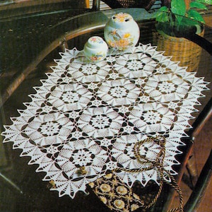 Puede incluir: Un tapete blanco de ganchillo con un patrón floral, colocado sobre una mesa de cristal. Dos tarros de cerámica decorativos con diseños florales están sobre el tapete. También se ve un pequeño bolso estampado con una correa dorada.