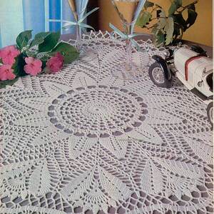 May include: White crocheted table runner with a circular design in the center. The runner is on a table with two glasses of liquid and a toy car.