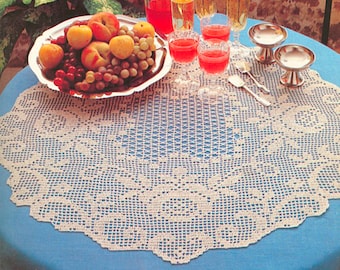 May include: A white crocheted doily with a delicate floral design sits on a blue tablecloth. A bowl of fruit and glasses of red juice are on the table.