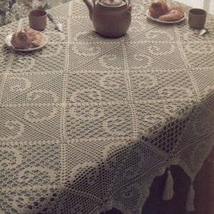 May include: A cream-colored, crocheted tablecloth with a repeating floral pattern. The tablecloth is draped over a table, with a teapot, cups, and plates of pastries on top. Tassels hang from the edges of the tablecloth.