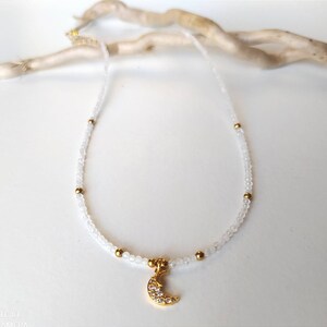 May include: A delicate white beaded necklace with a gold crescent moon charm. The moon charm is adorned with small sparkling stones.