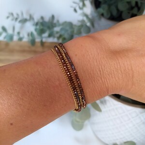 May include: A set of three bracelets featuring a gold chain and a brown beaded bracelet. The bracelets are stacked on a person's wrist.