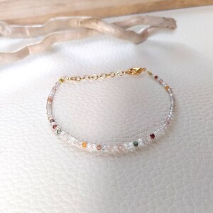 May include: A delicate gold chain bracelet with small, colorful gemstone beads. The beads are a mix of clear, pink, orange, green, and red.